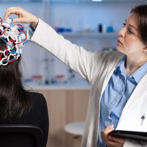 Back view of woman patient wearing performant eeg headset sitting on chair in neurological research laboratory while medical researcher adjusting it, examining nervous system typing on tablet.
